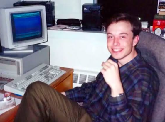 A young Elon Musk in a plaid shirt sits beside a beige CRT computer and keyboard